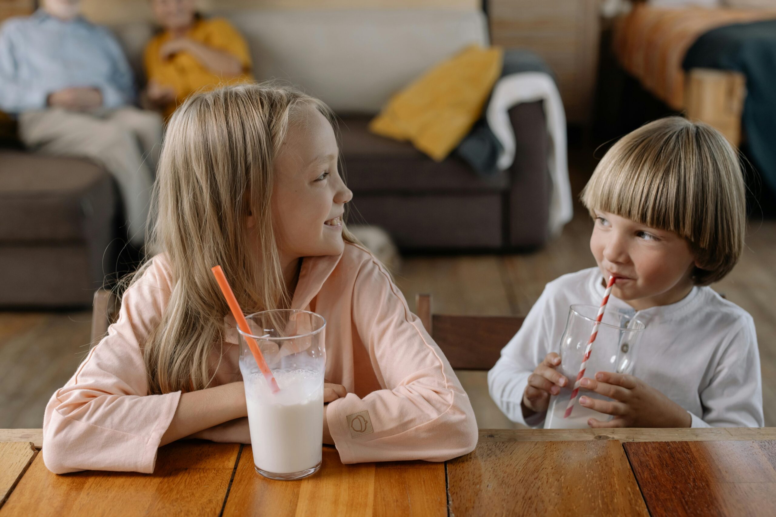 Two children smiling and drinking milk with straws indoors, enjoying each other's company.