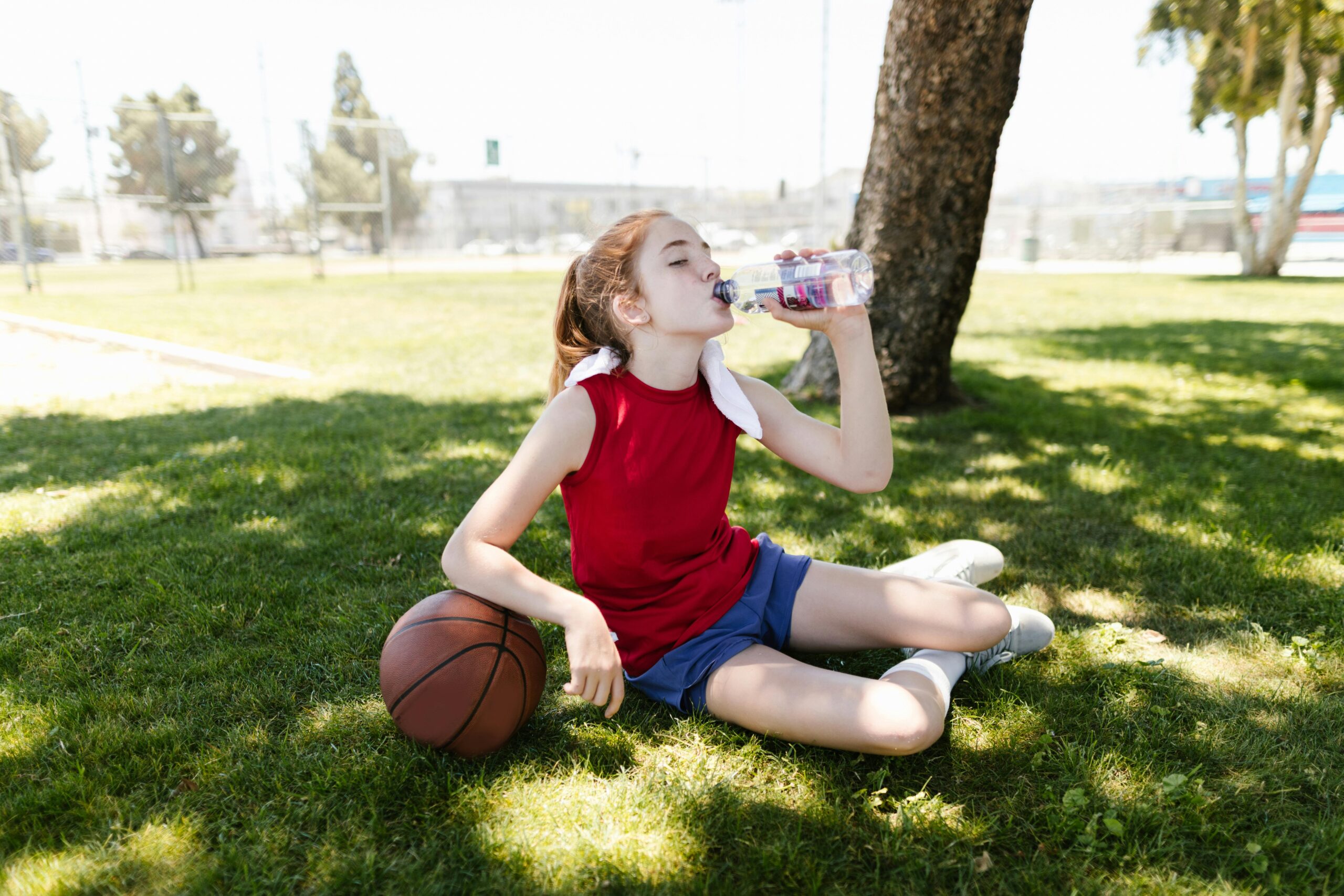 A young girl in sportswear enjoys a refreshing drink in the park after playing basketball.