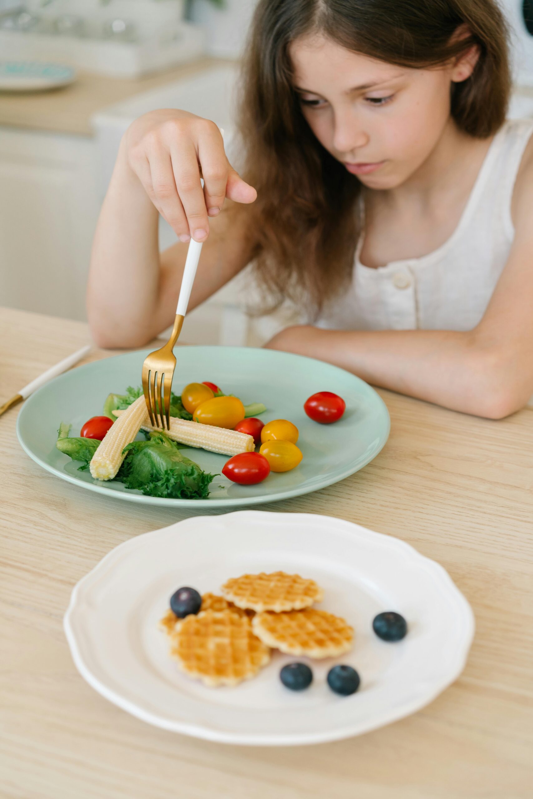 A young girl in a white dress enjoys a healthy salad with fresh vegetables on a plate.