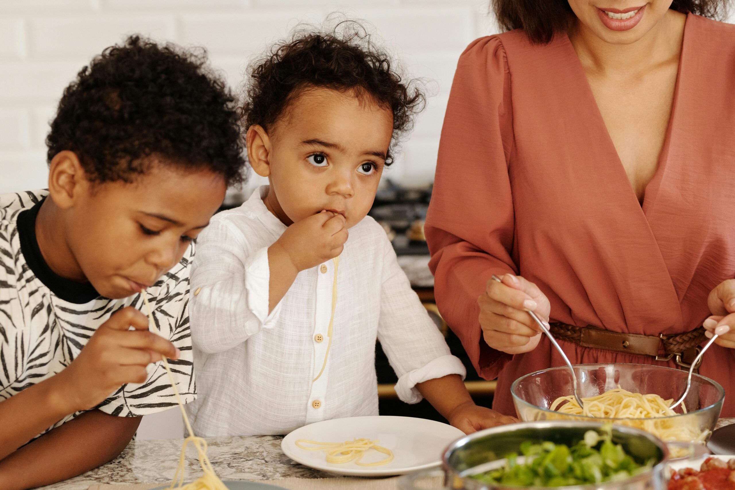 Mother and children eating spaghetti indoors at home kitchen.