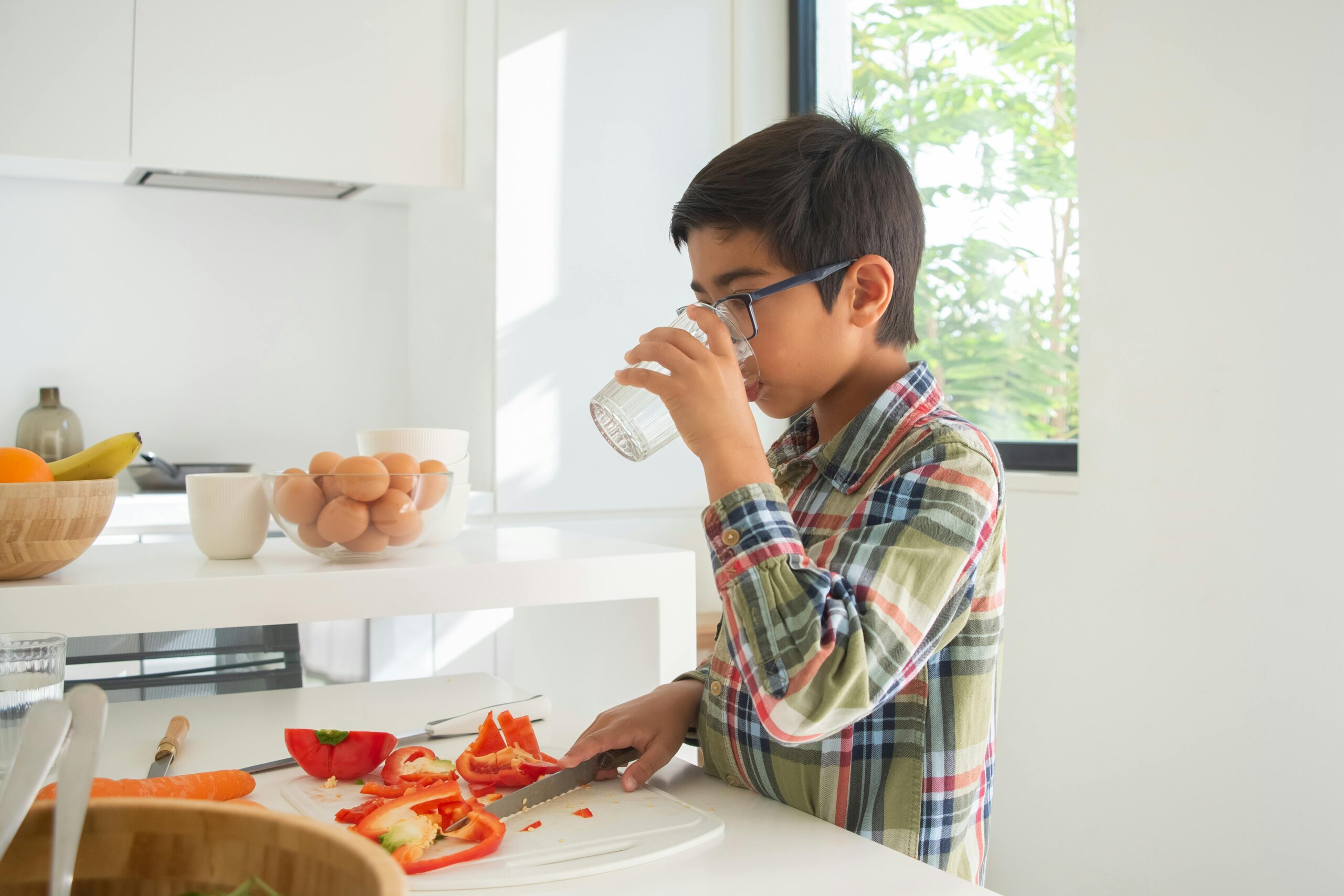 Young boy with glasses drinks water in a bright kitchen while preparing a healthy snack.
